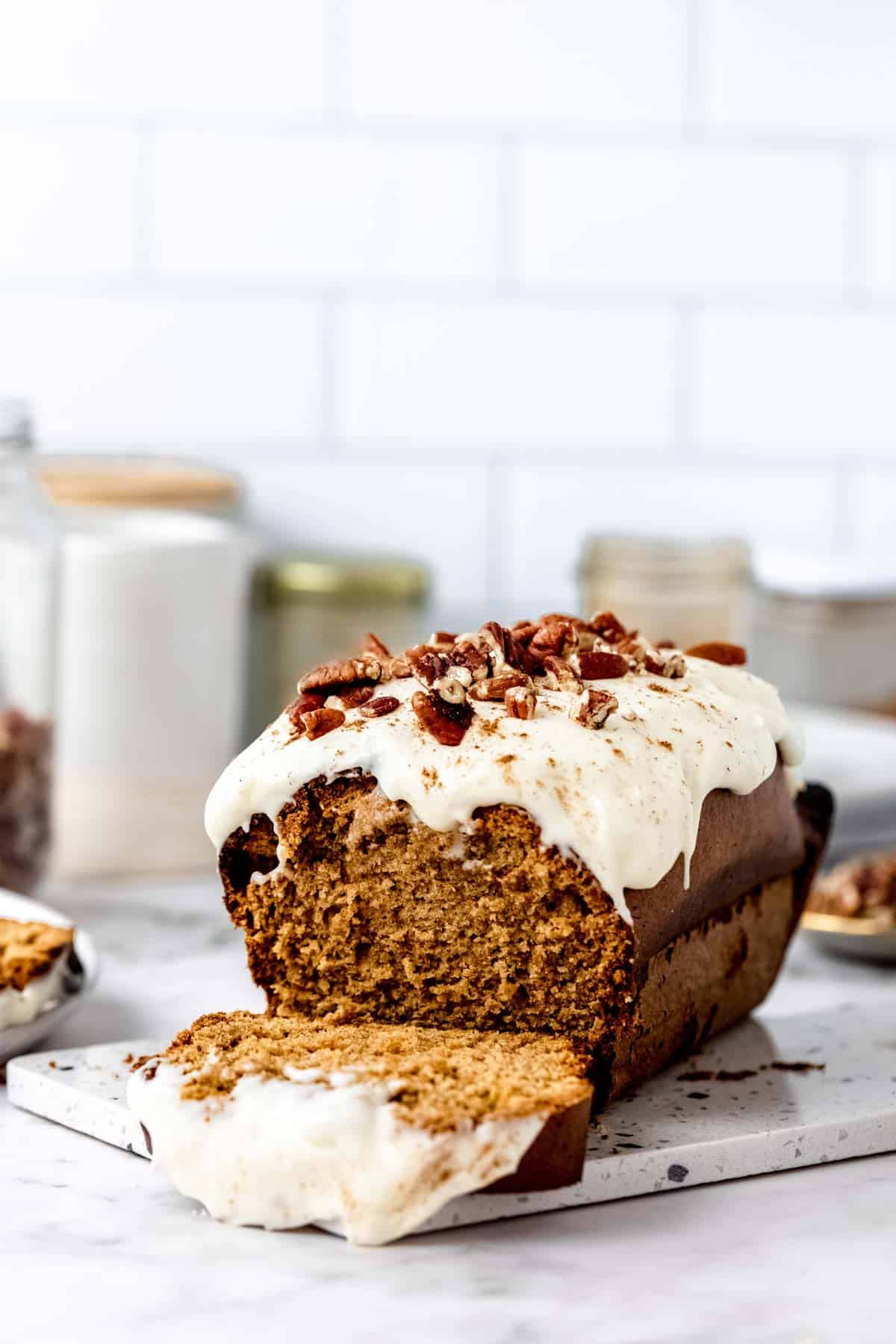 A gluten-free gingerbread loaf frosted with cream cheese icing and topped with pecan nuts, sliced on a white countertop.