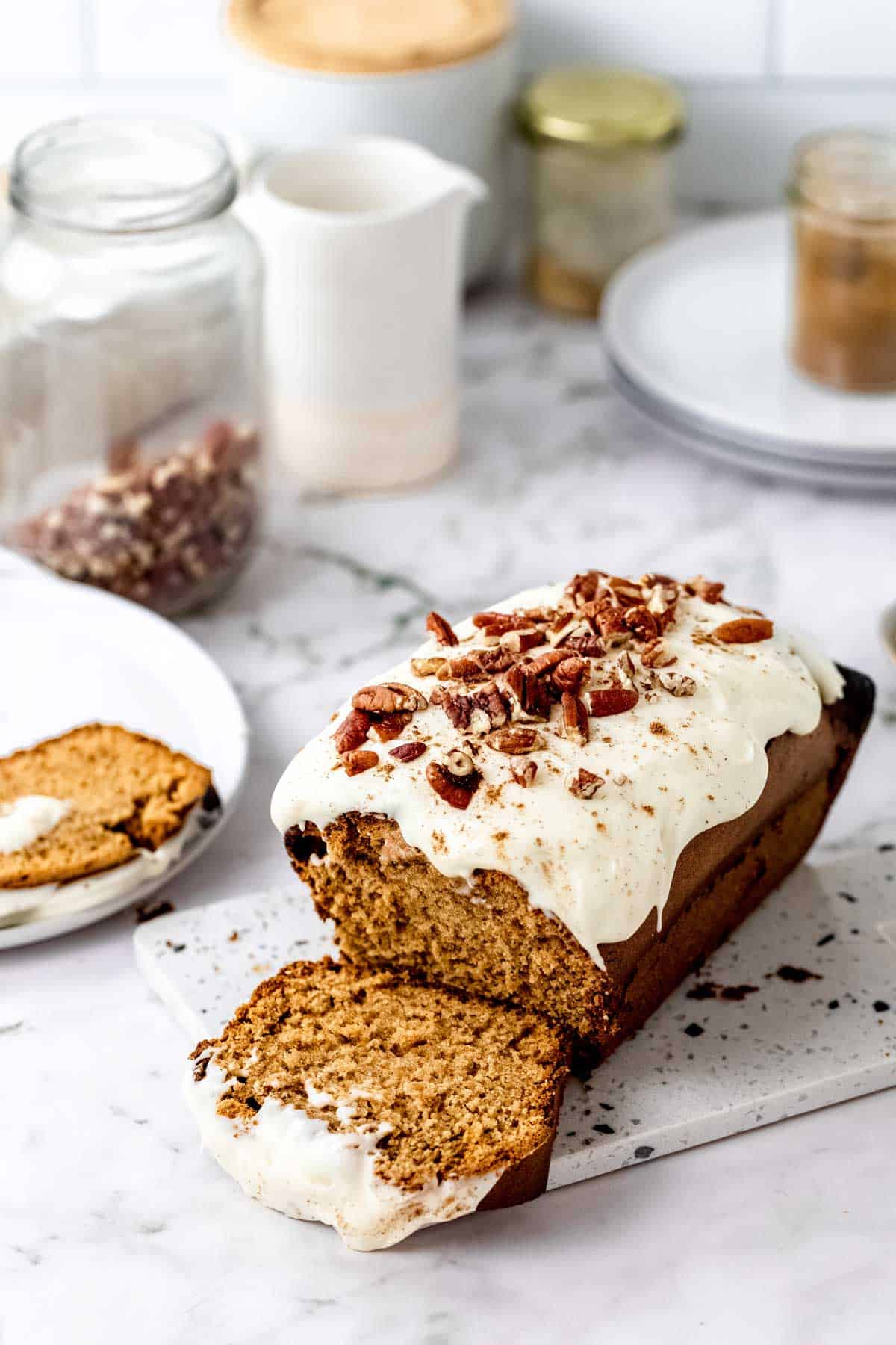 A gluten-free gingerbread loaf frosted with cream cheese icing and topped with pecan nuts, sliced on a white countertop.