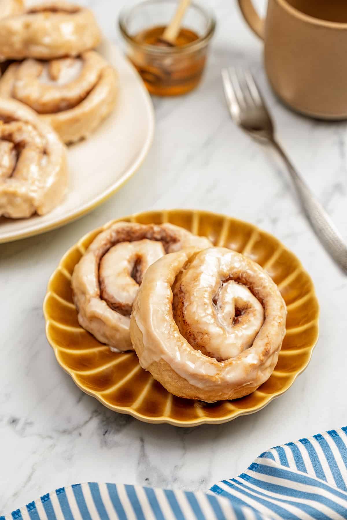 Two gluten-free honey buns on a brown plate next to a plate of honey buns.