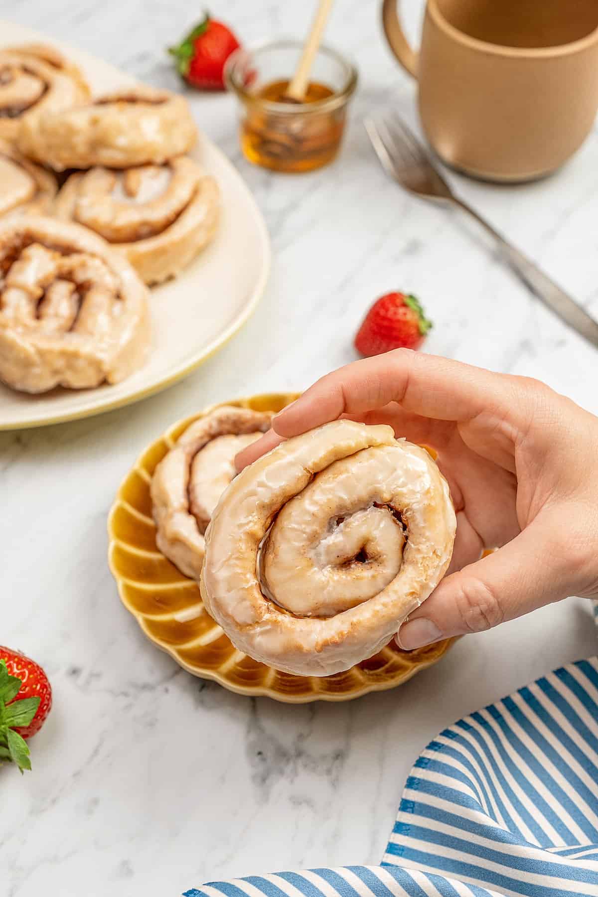 A hand holding a honey bun over a plate, with more buns in the background.