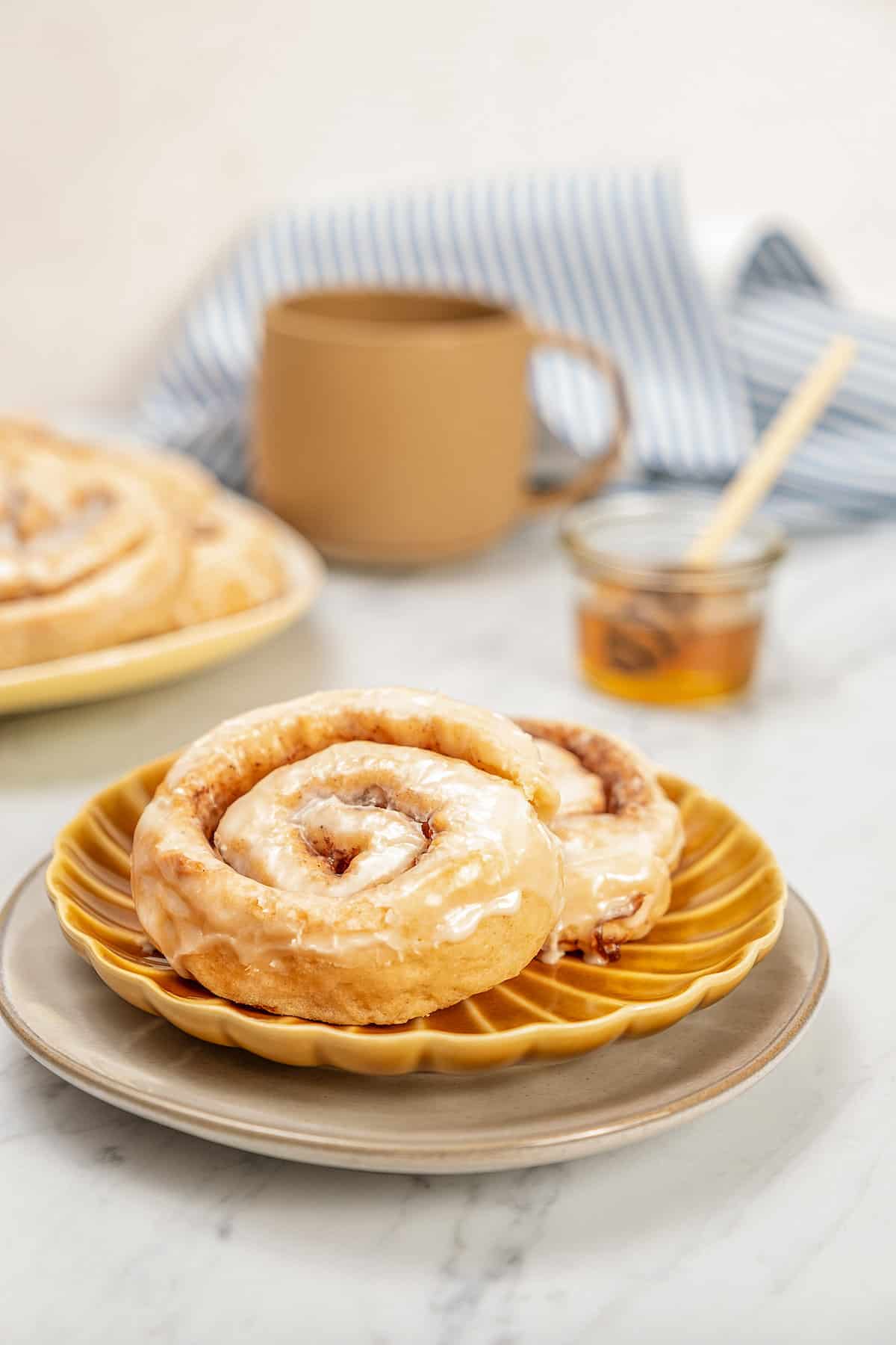 Two gluten-free honey buns on a brown plate with a coffee mug and jar of honey in the background.