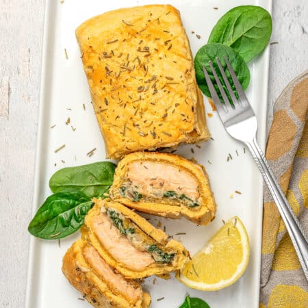 Gluten-free Salmon Wellington cut into slices on a platter, next to a lemon wedge and fresh spinach leaves.