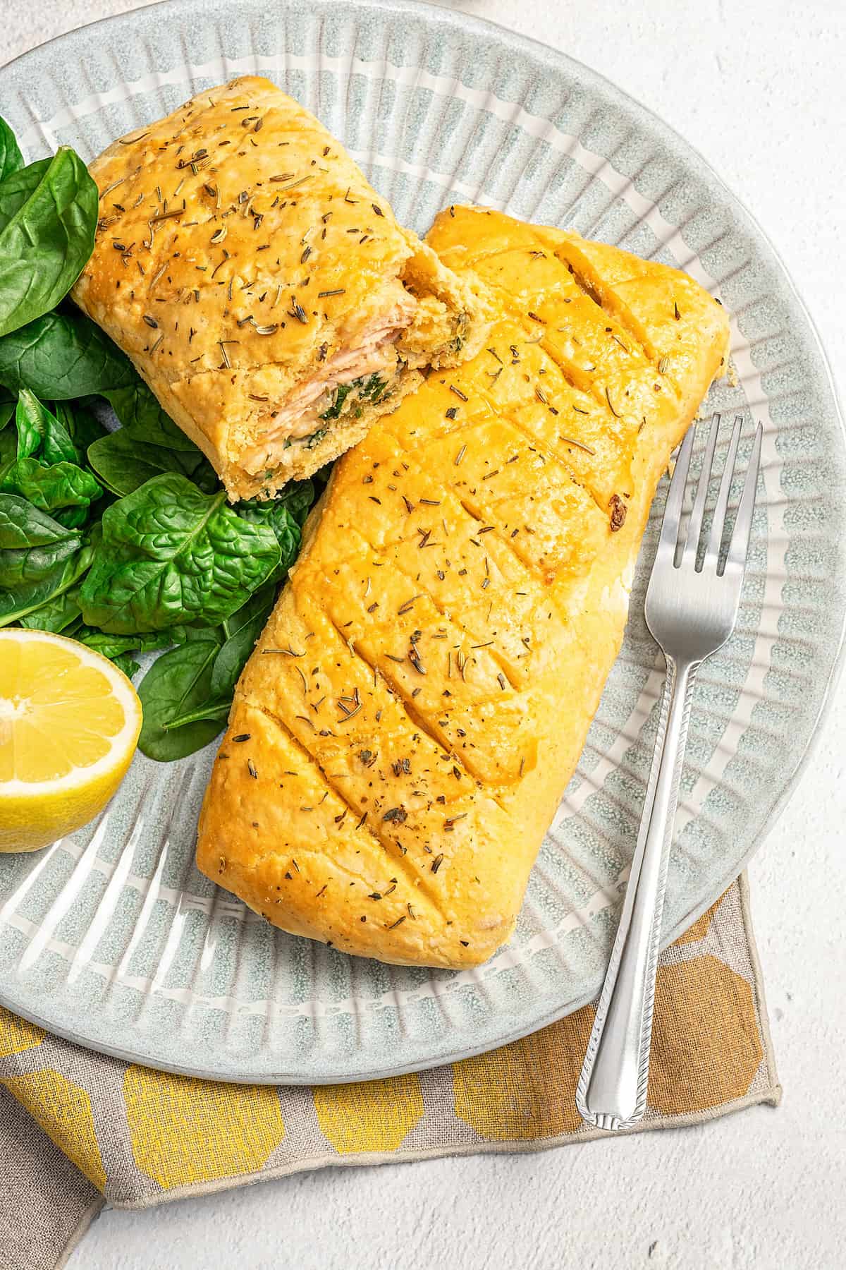 A whole gluten-free Salmon Wellington plus a half Wellington on a platter, next to a lemon wedge and fresh spinach leaves and a fork.