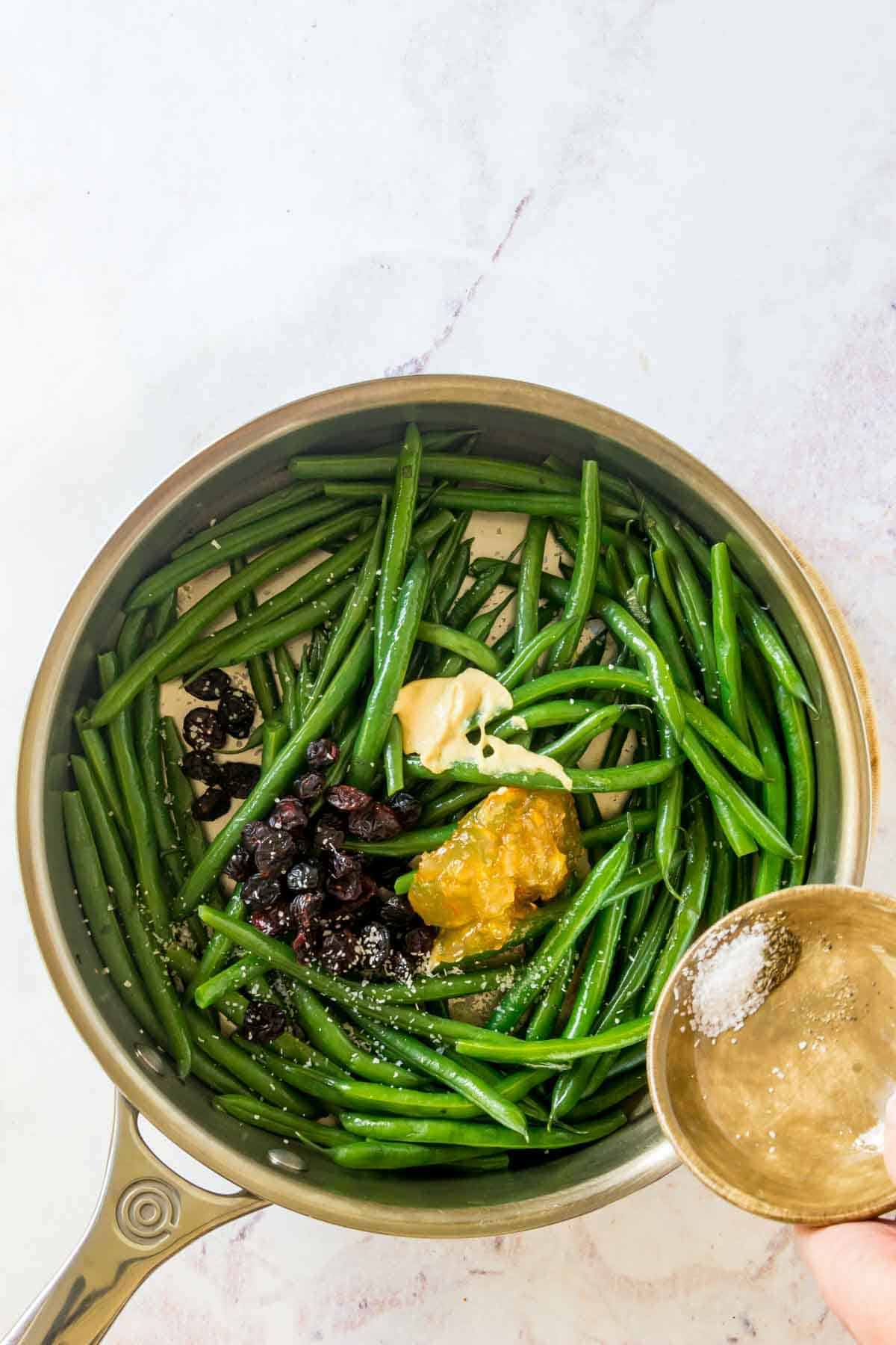 Salt and pepper bring sprinkled on a green beans in a skillet that have Dijon mustard, orange marmalade, and dried cranberries on top.