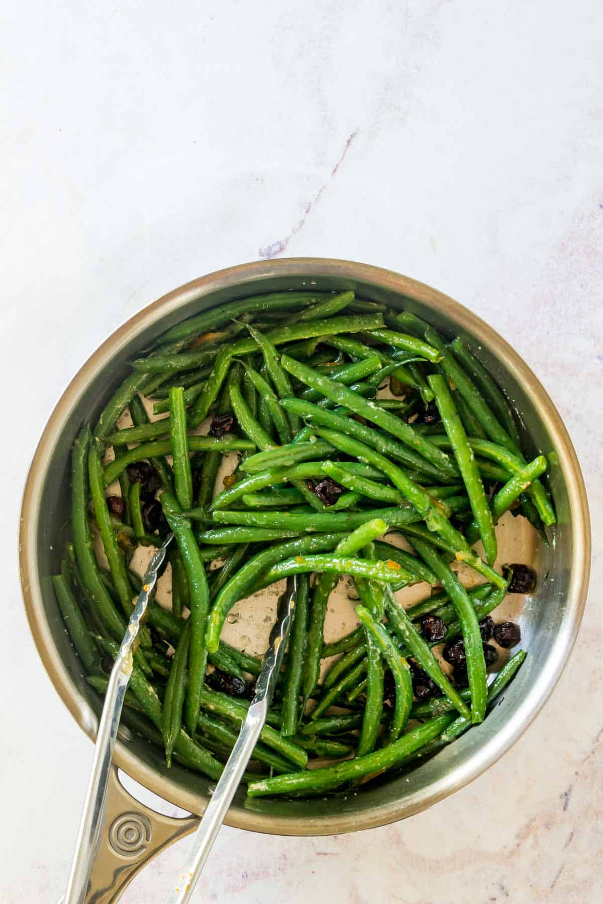 Glazed green beans with dried cranberries being tossed together with tongs in a skillet.