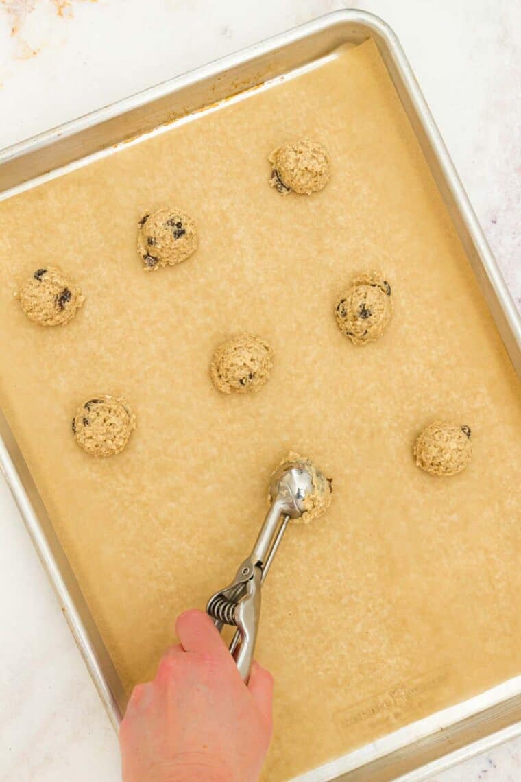 A hand uses a cookie scoop to portion out gluten-free oatmeal raisin cookie dough onto a baking sheet.