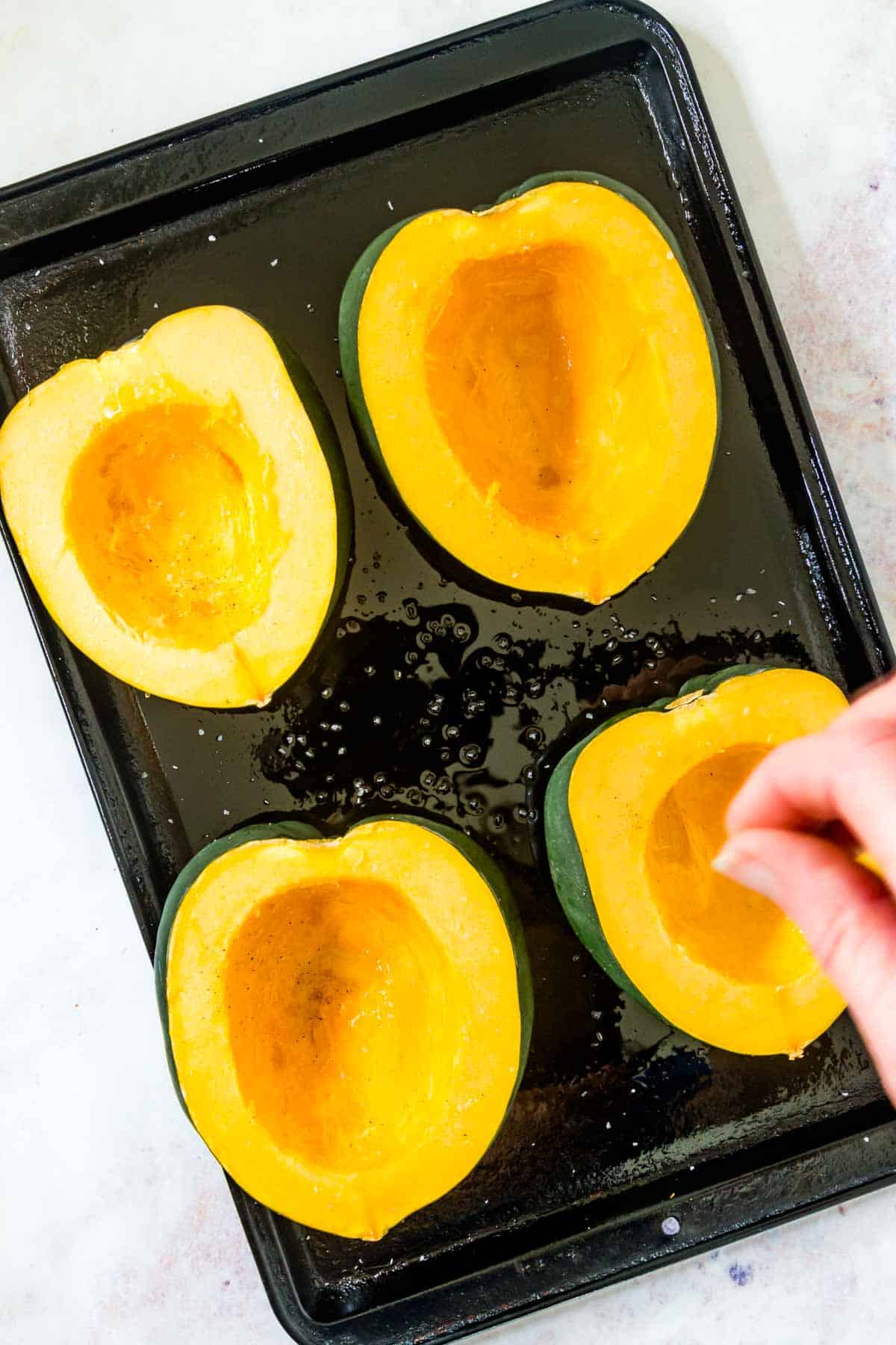 Four acorn squash halves cut side up on a baking sheet with pepper being sprinkled on them.