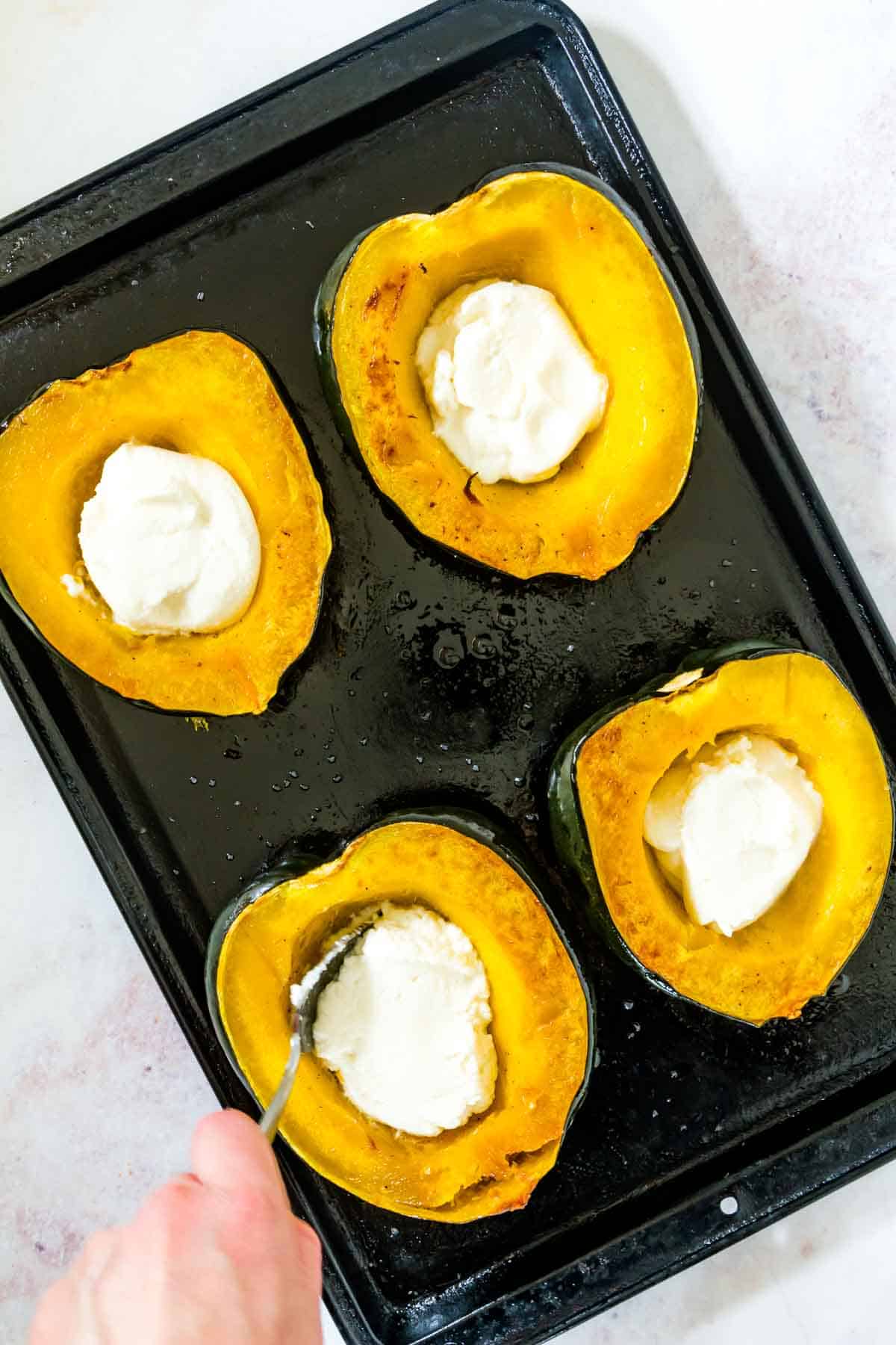 Ricotta cheese being spooned into the roasted acorn squash halves on a sheet pan.
