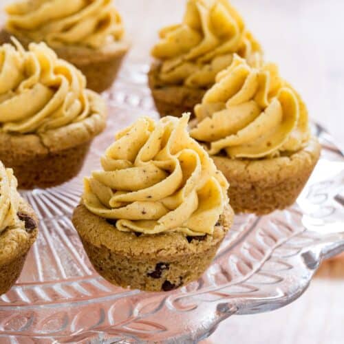 Several chocolate chip cookie cups with a swirl of pumpkin frosting on a glass cake stand.