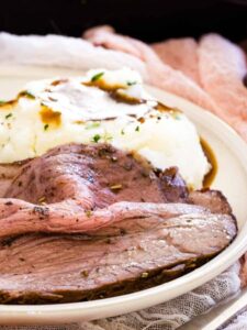 Slices of bottom round roast beef on a plate with mashed potatoes and green beans.