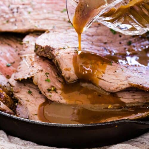 Brown gravy being poured over slices of bottom round roast beef in a cast iron skillet.