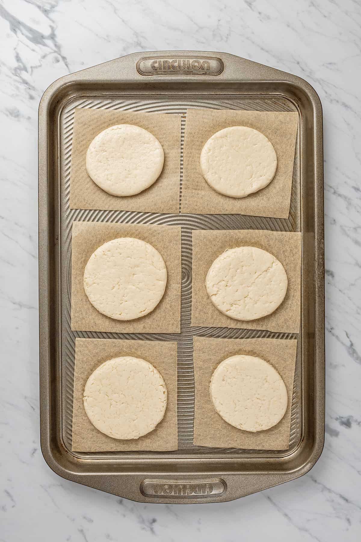 Six round discs of bao bun dough on squares of parchment paper on a baking sheet.