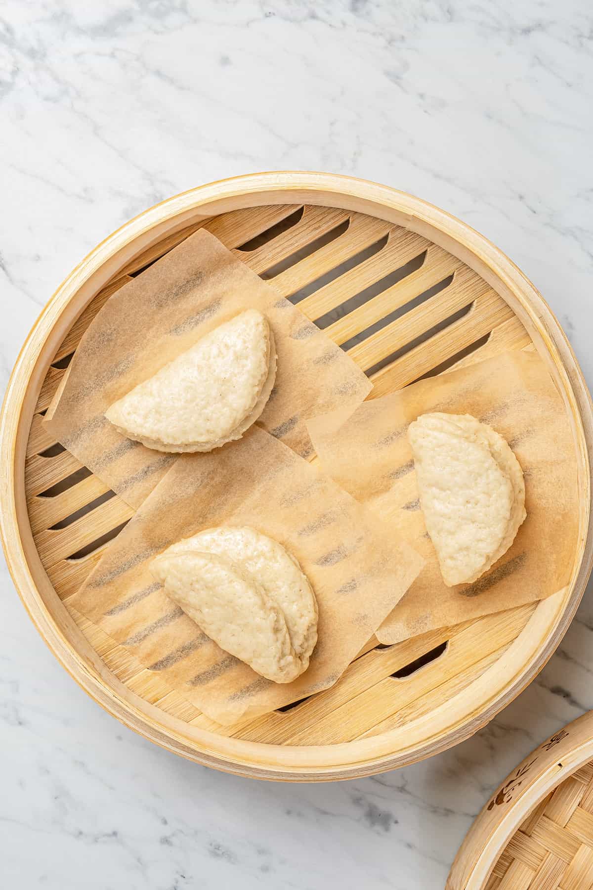 Three gluten-free steamed bao buns on squares of parchment paper inside a bamboo steamer basket.