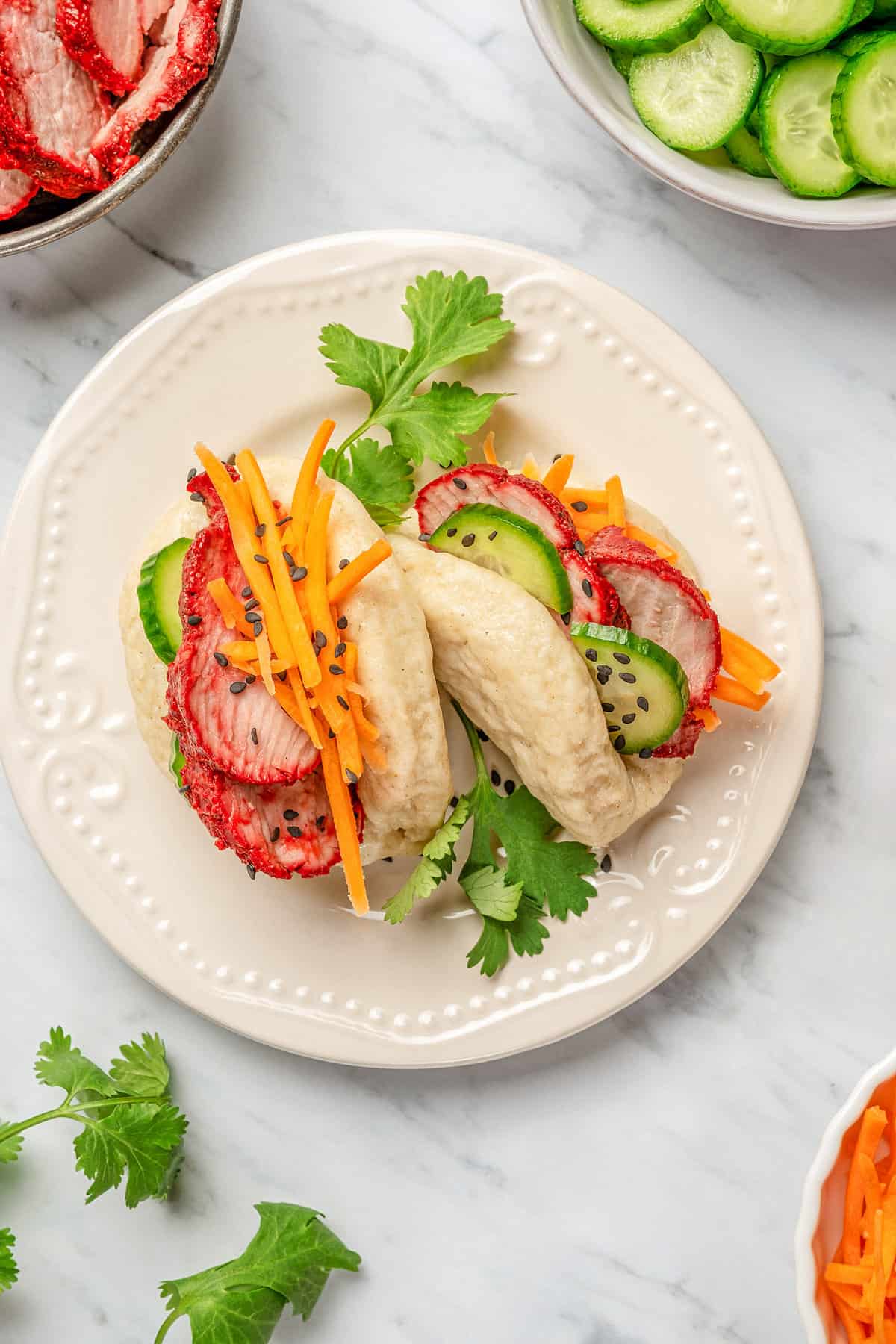 Overhead view of two gluten-free bao buns filled with char siu pork and veggies on a plate, surrounded by bowls of toppings..