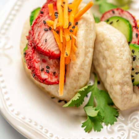Two gluten-free bao buns filled with char siu pork and veggies on a plate, garnished with coriander leaves.