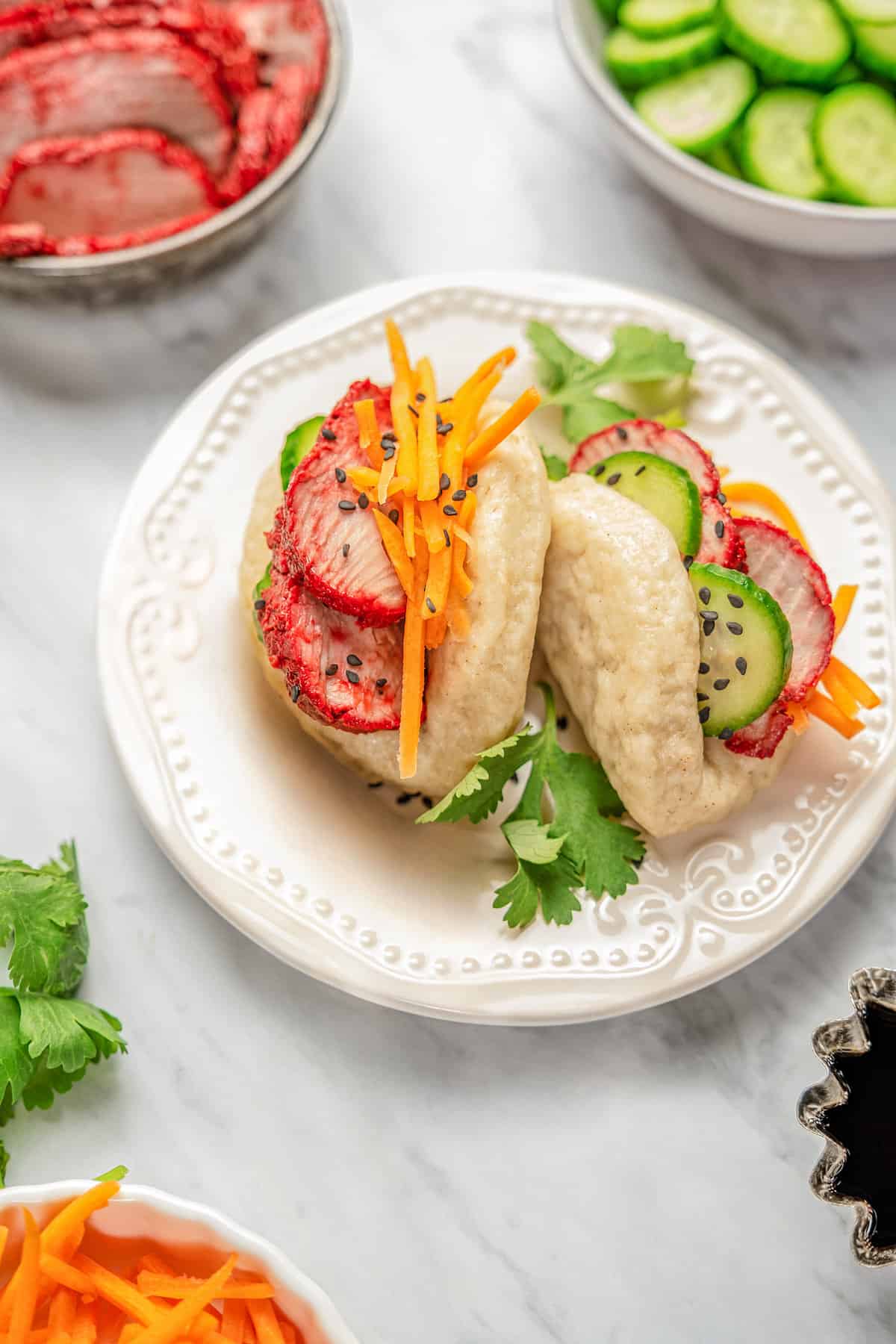 Overhead view of two gluten-free bao buns filled with char siu pork and veggies on a plate, surrounded by bowls of toppings..