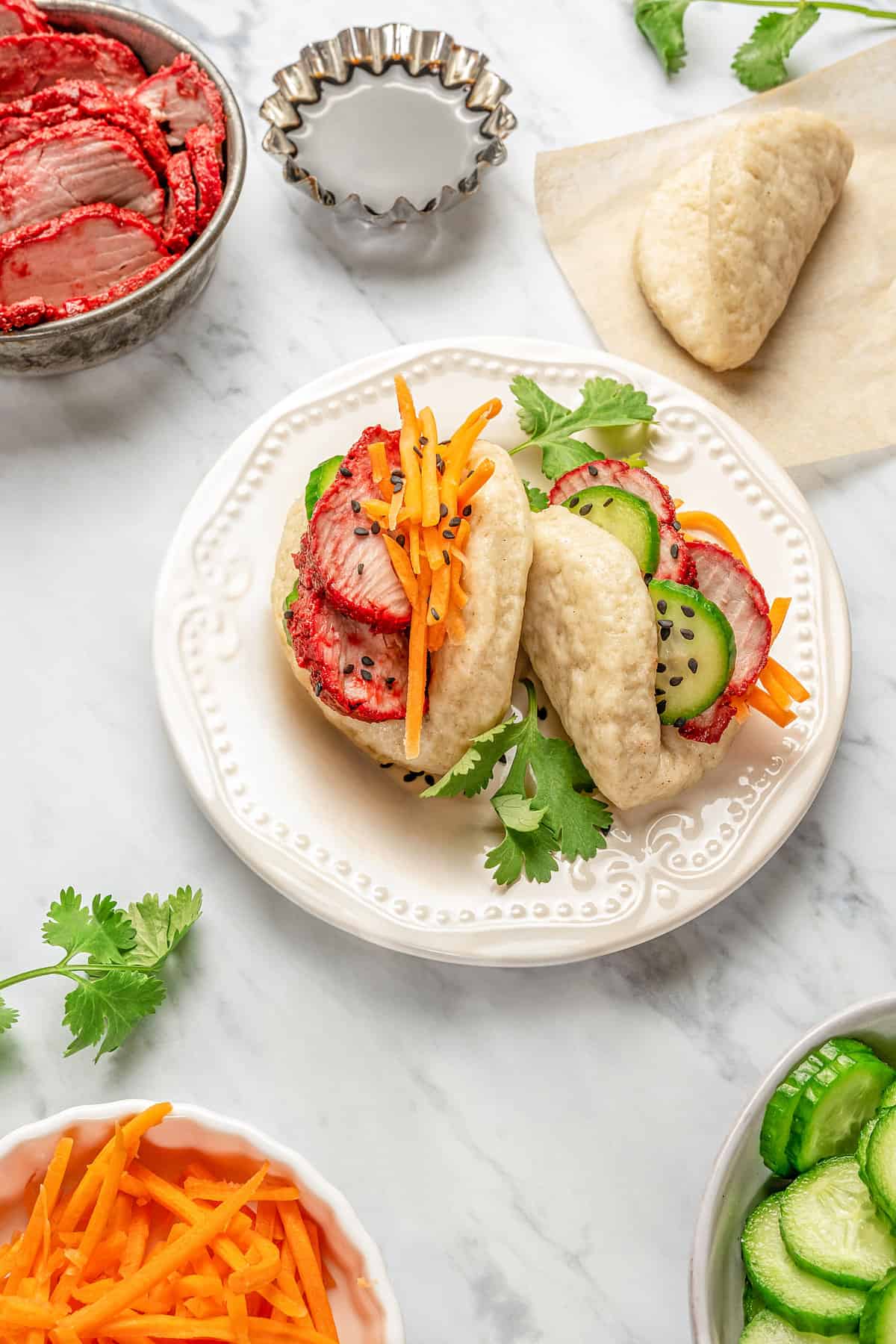 Overhead view of two gluten-free bao buns filled with char siu pork and veggies on a plate, surrounded by bowls of toppings..