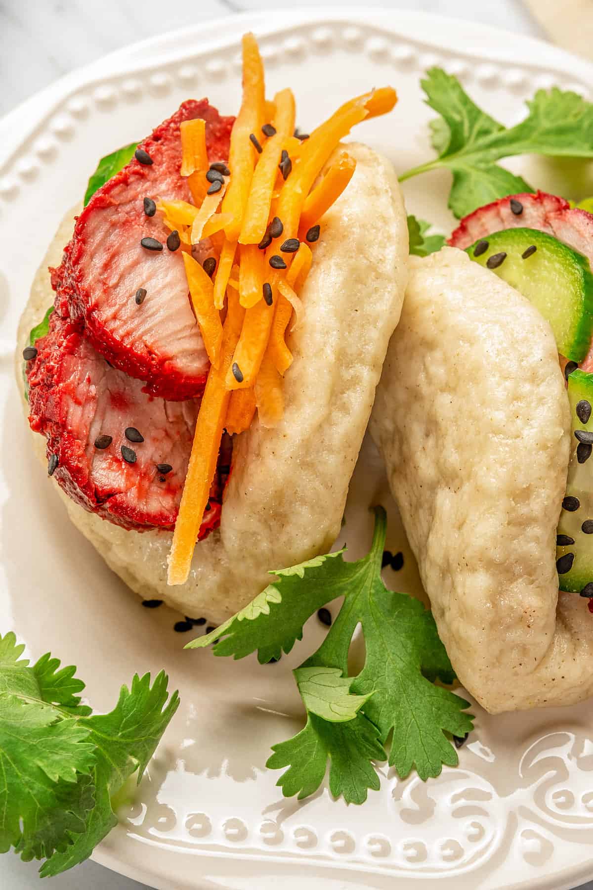 Close up of two gluten-free bao buns filled with char siu pork and veggies on a plate, garnished with coriander leaves.