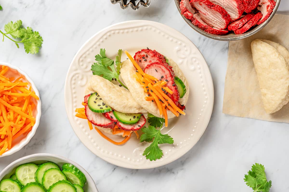 Overhead view of two gluten-free bao buns filled with char siu pork and veggies on a plate, surrounded by bowls of toppings..