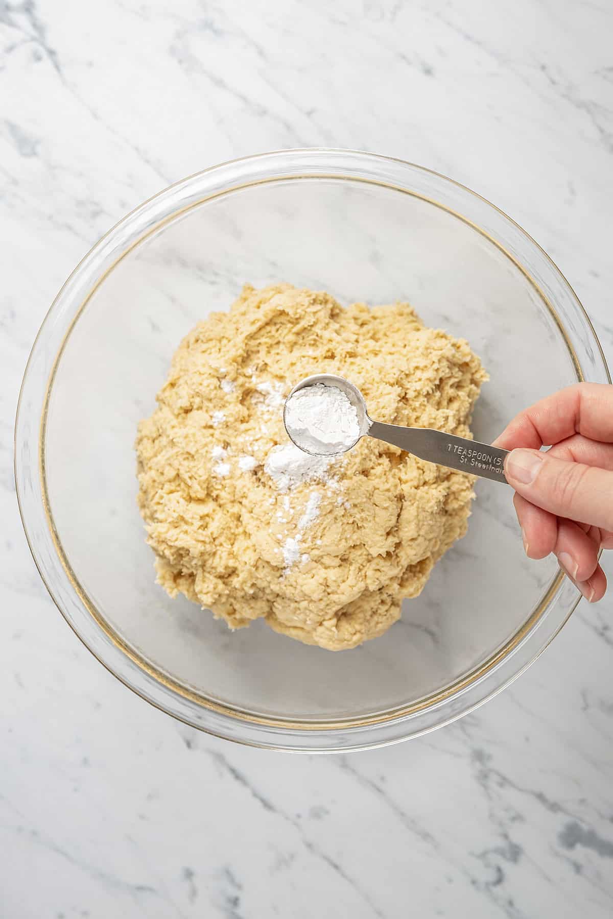 A hand holding a teaspoon sprinkles baking soda over proofed gluten-free bread dough in a glass bowl.