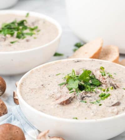 Bowls of cream of mushroom soup garnished with fresh parsley, next to slices of crusty bread and mushrooms.