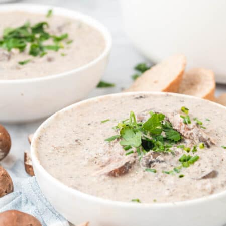 Bowls of cream of mushroom soup garnished with fresh parsley, next to slices of crusty bread and mushrooms.