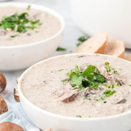 Bowls of cream of mushroom soup garnished with fresh parsley, next to slices of crusty bread and mushrooms.