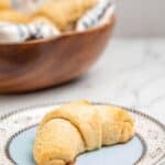 A gluten free crescent roll on a china plate with a bowl of more rolls in the background.