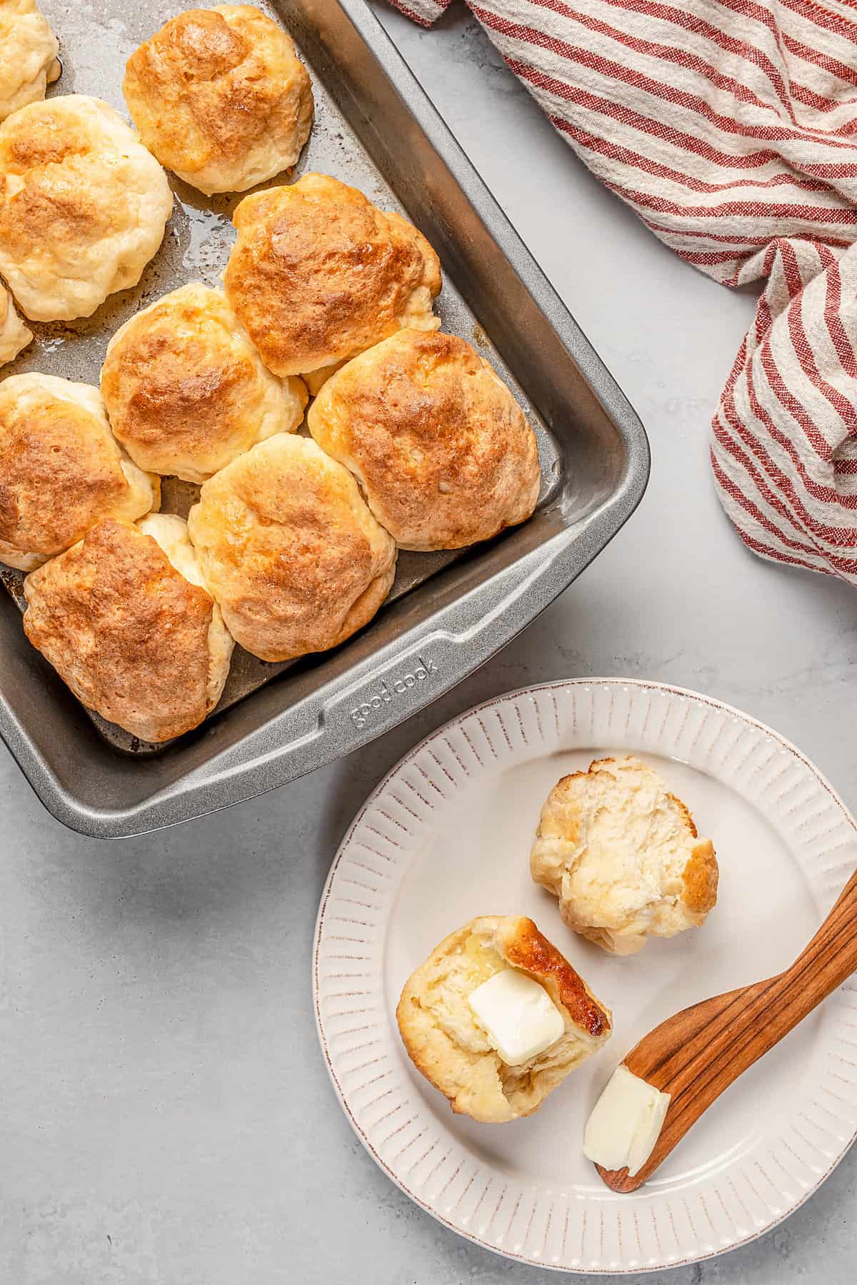 A gluten-free Hawaiian roll cut in half on a plate, topped with butter, next to more rolls in a baking pan.