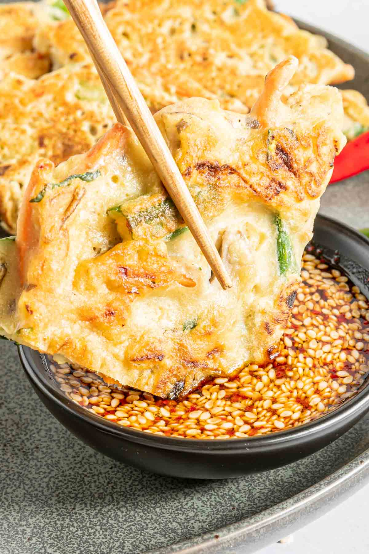 Chopsticks holding a vegetable pancake being dipped in a bowl of Korean dipping sauce.