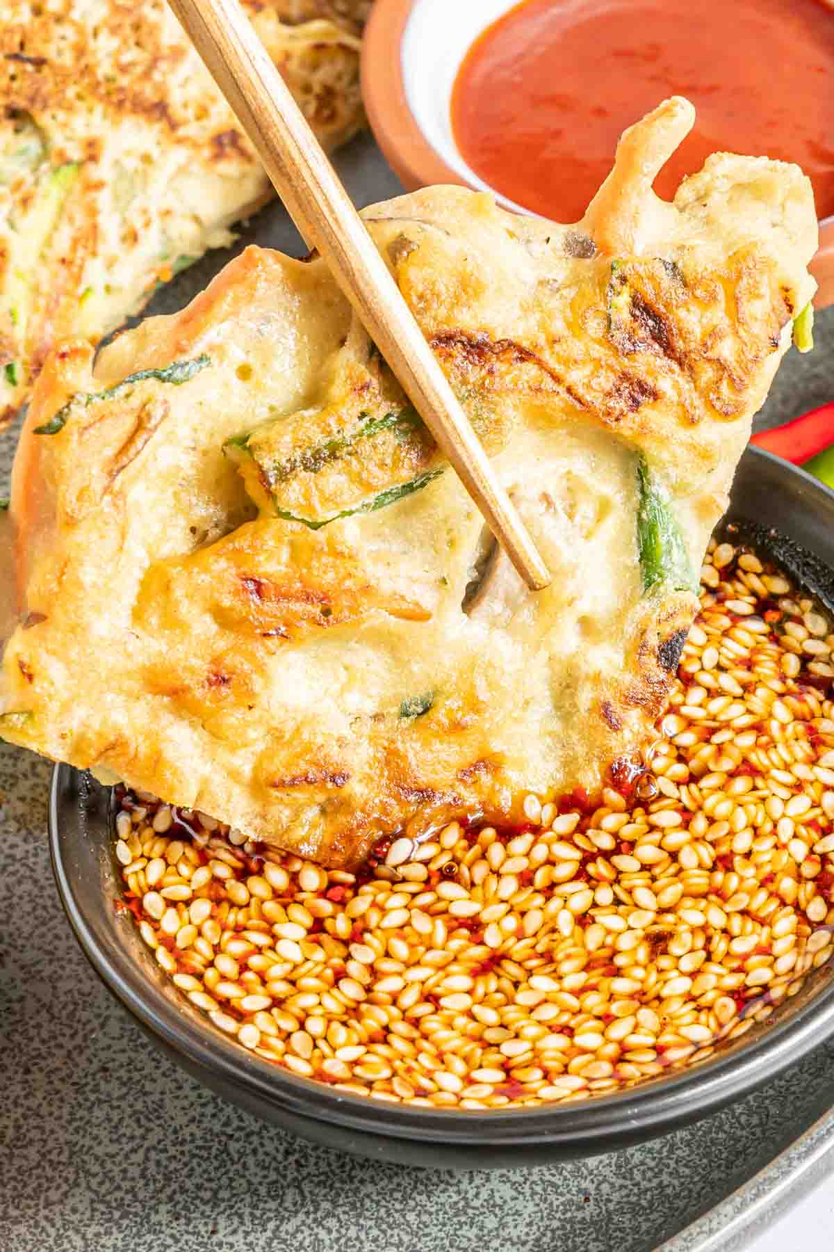 Chopsticks holding a vegetable pancake being dipped in a bowl of Korean dipping sauce.