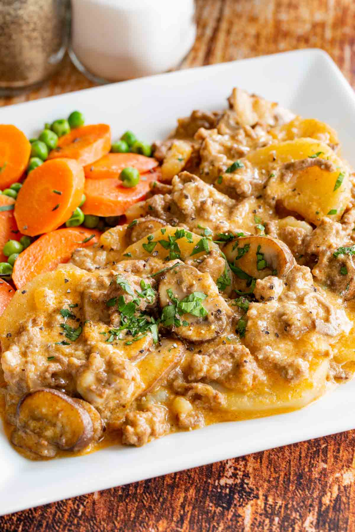 Ground beef and potatoes with mushrooms in a creamy sauce served on a white plate with peas and carrots.