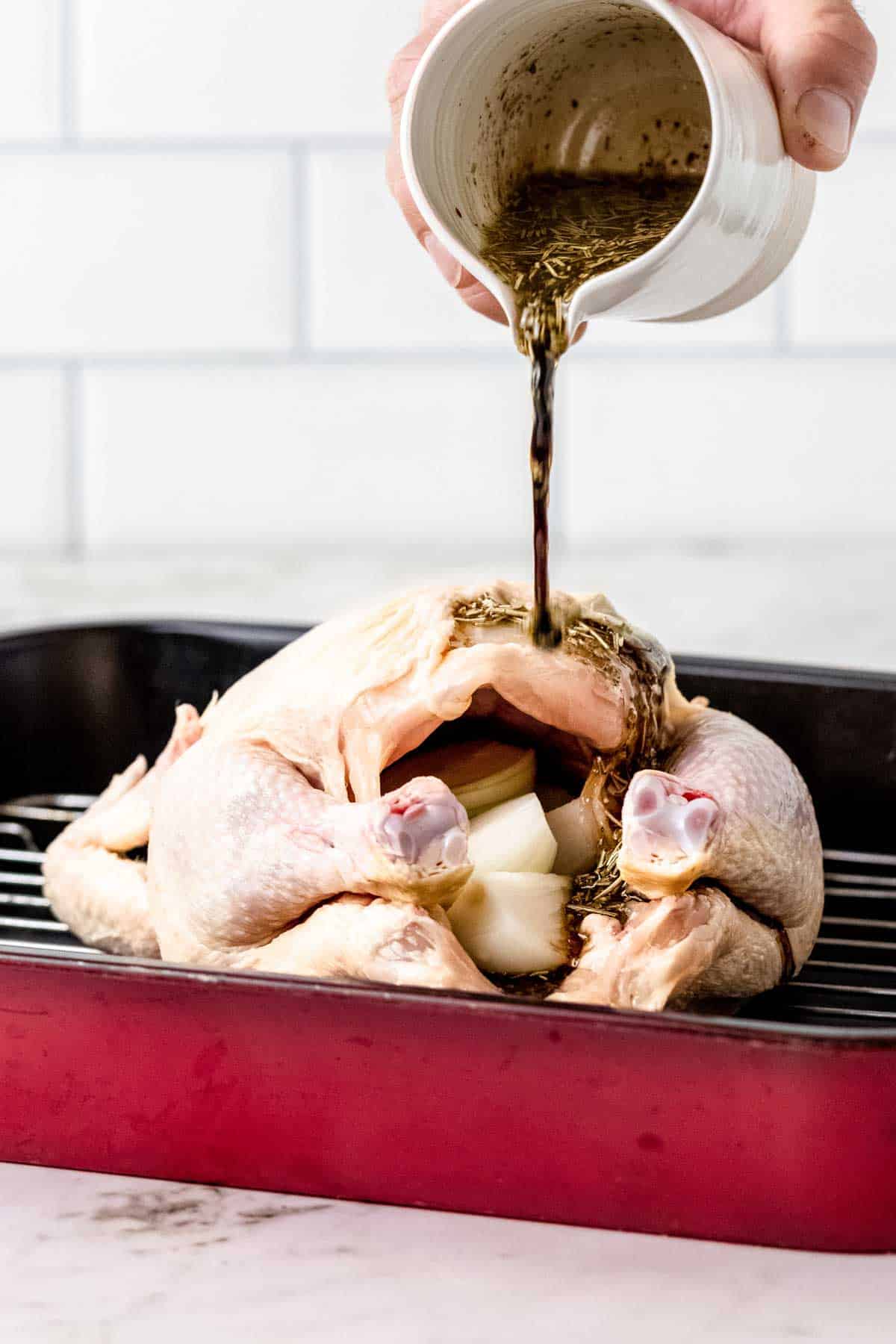 Rosemary balsamic marinade is poured over a whole raw chicken stuffed with chopped onions in a red roasting pan.