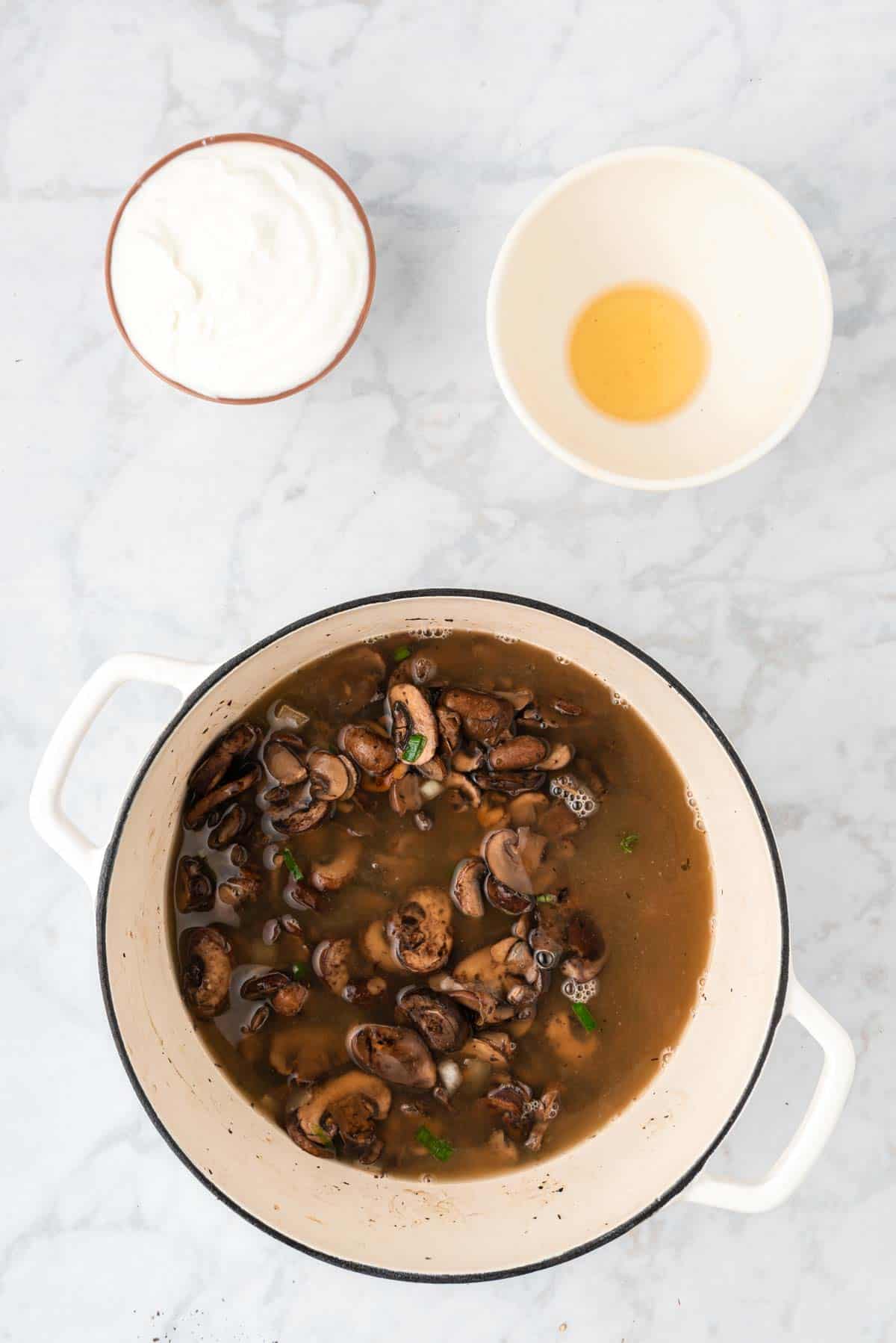 Sauteed mushrooms and broth in a large pot next to a bowl of Greek yogurt.