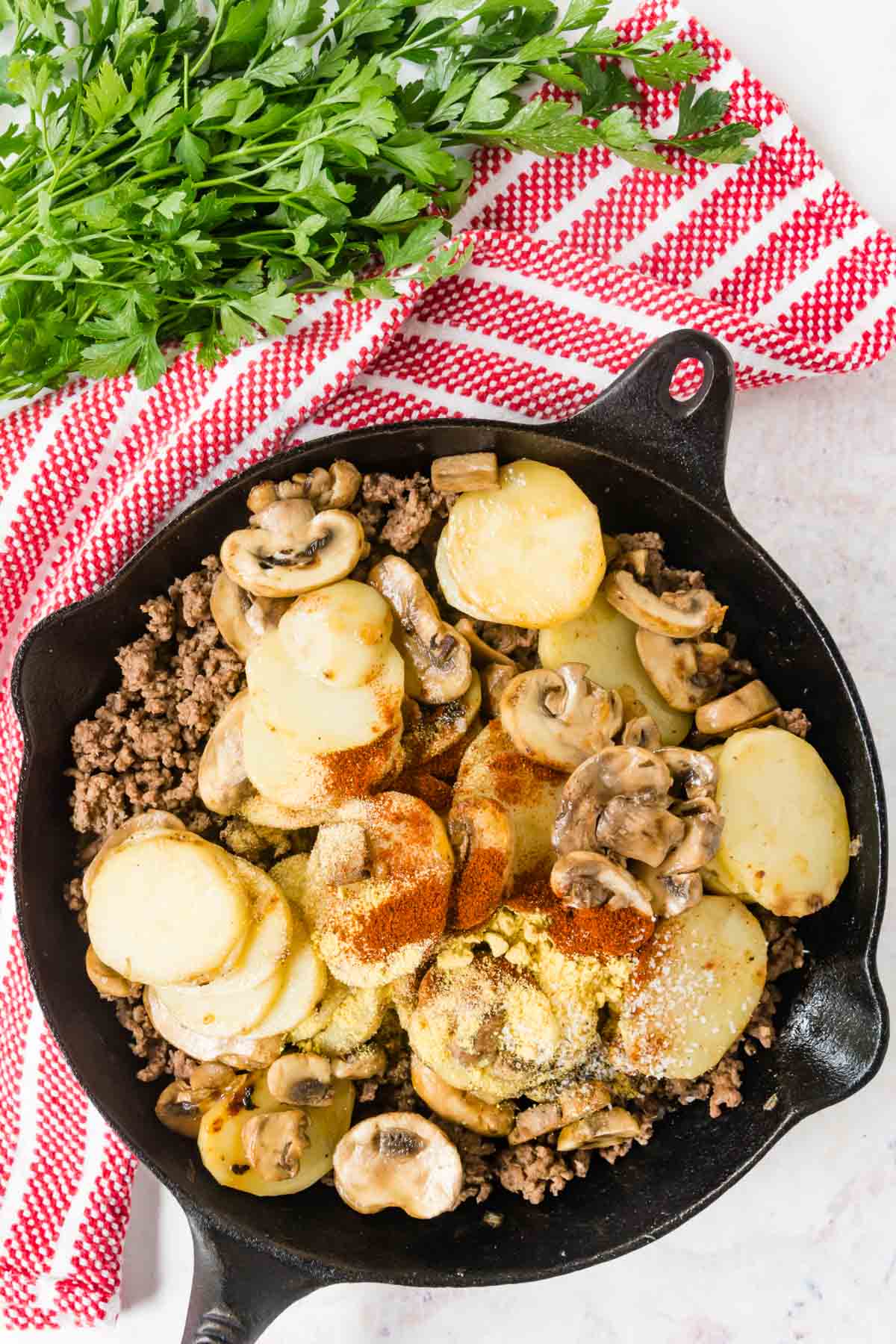 Spices sprinkled on top of meat, potatoes, and mushrooms in the skillet.