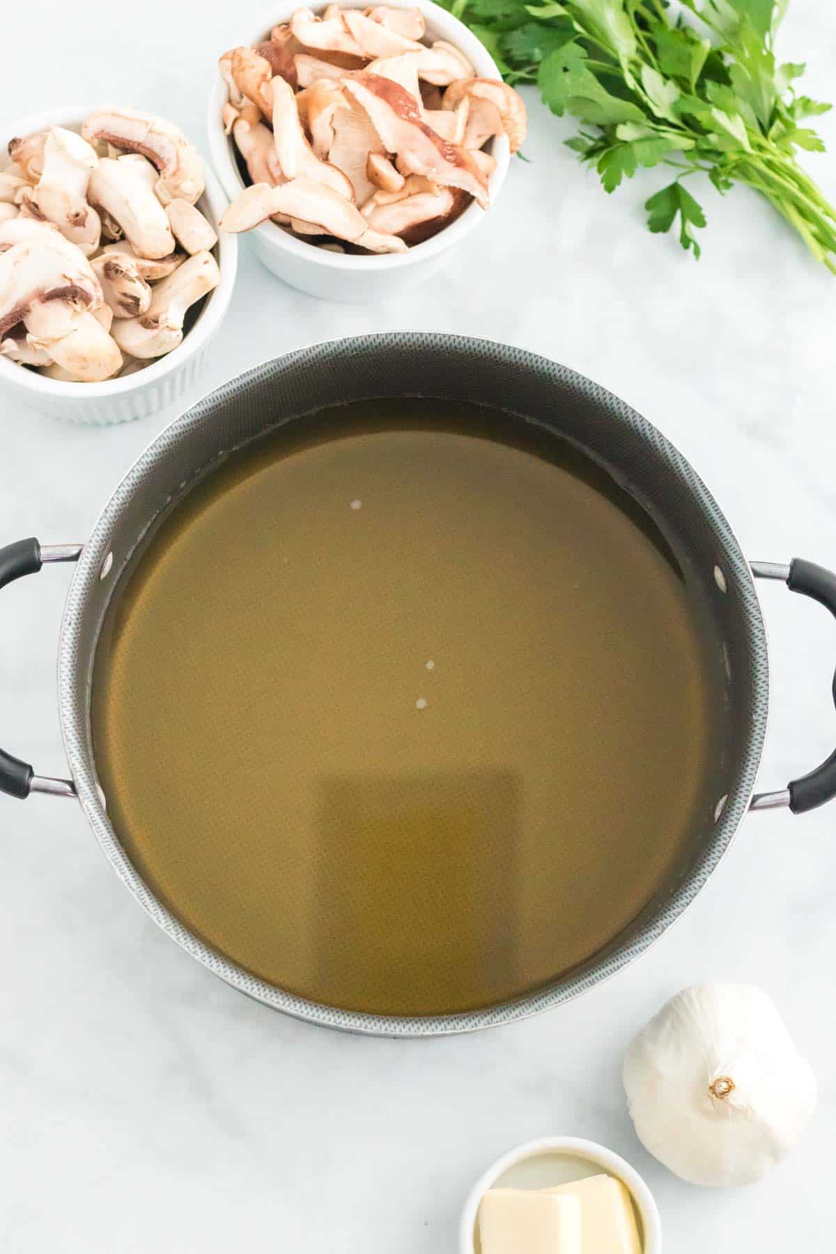 Overhead view of a pot of chicken stock next to bowls of chopped mushrooms, a bulb of garlic, and butter..