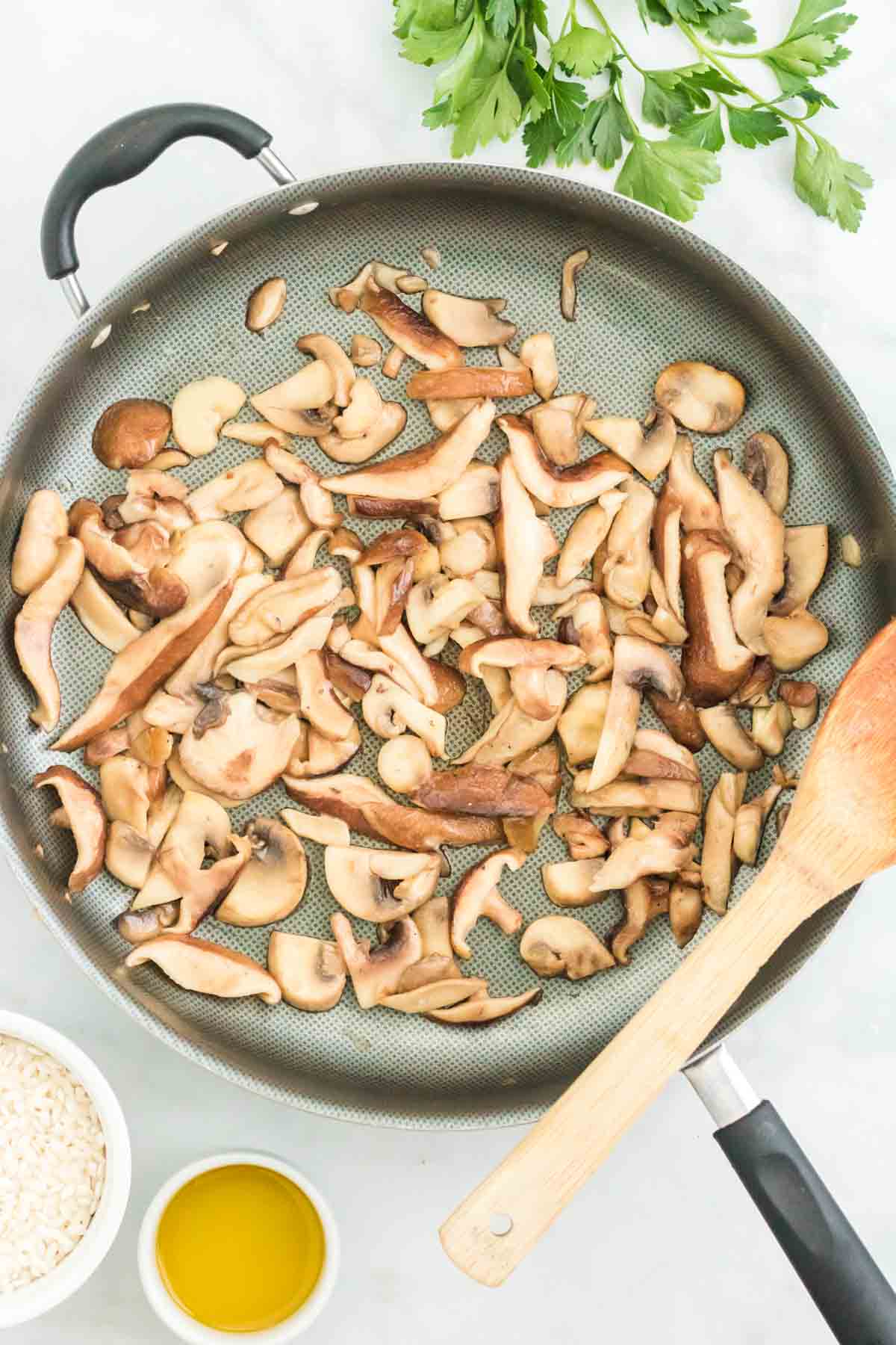 Sliced mushrooms sauteeing in a skillet next to a wooden spoon.