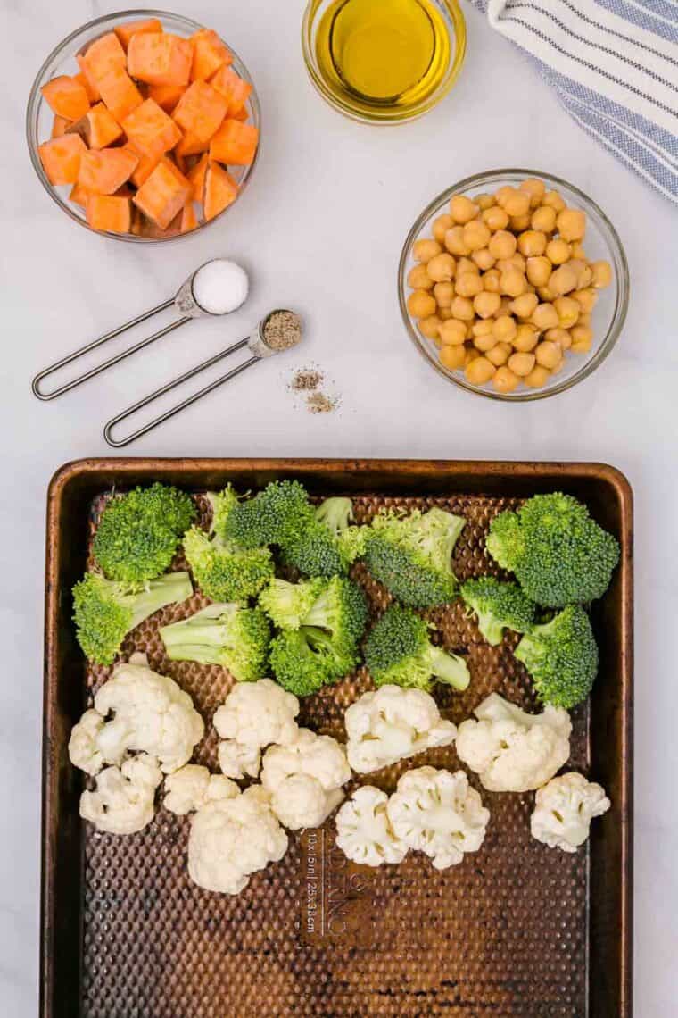 Broccoli and cauliflower florets on a baking sheet next to bowls of diced sweet potatoes and chickpeas.