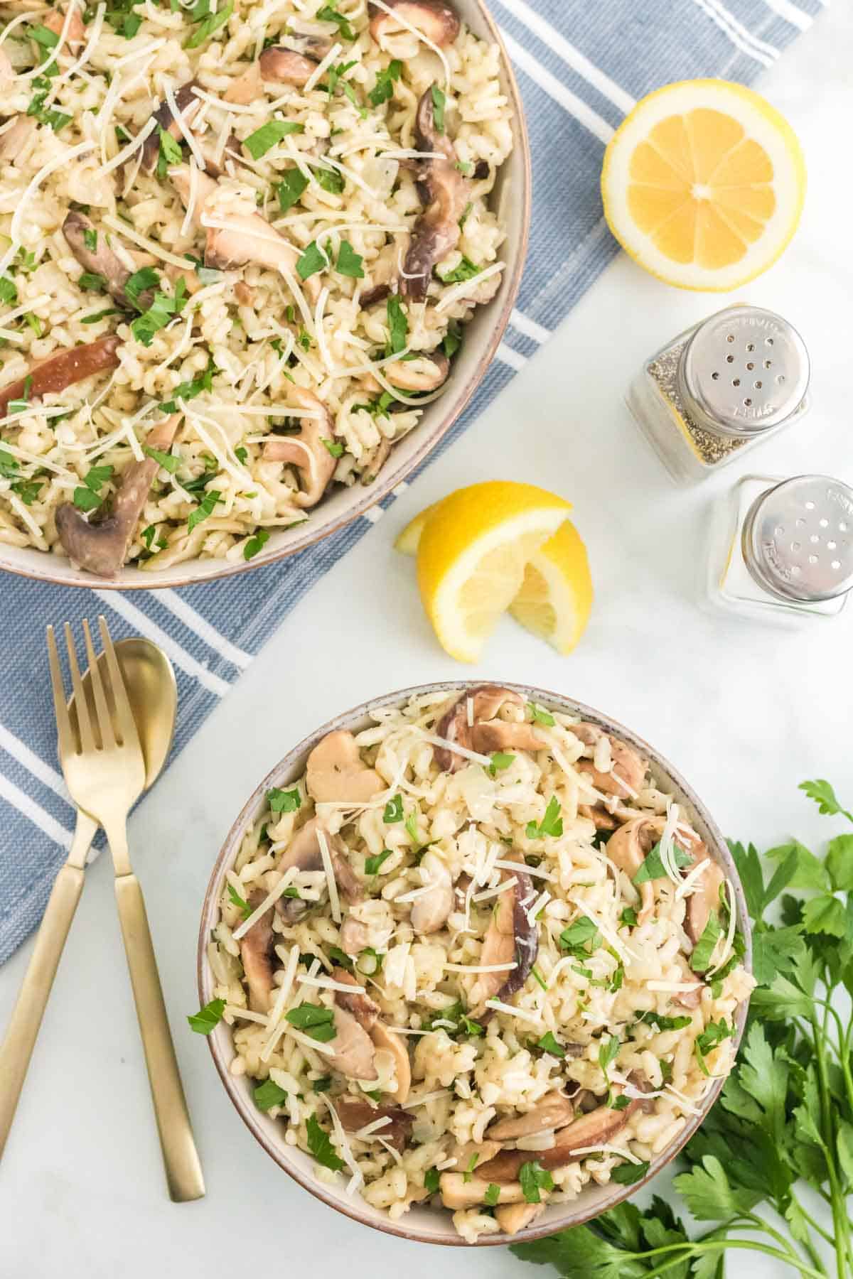 A bowl of mushroom risotto next to a skillet of risotto, lemon wedges, and salt and pepper shakers.