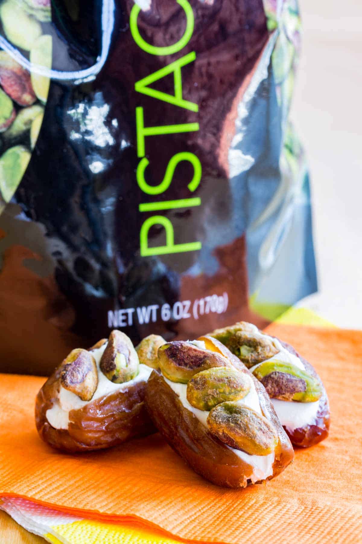 Three dates stuffed with cream cheese and pistachios on an orange napkin in front of a bag of pistachios.
