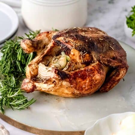 A whole rosemary balsamic roast chicken on a marble cutting board, next to a bowl of onions and fresh rosemary sprigs.