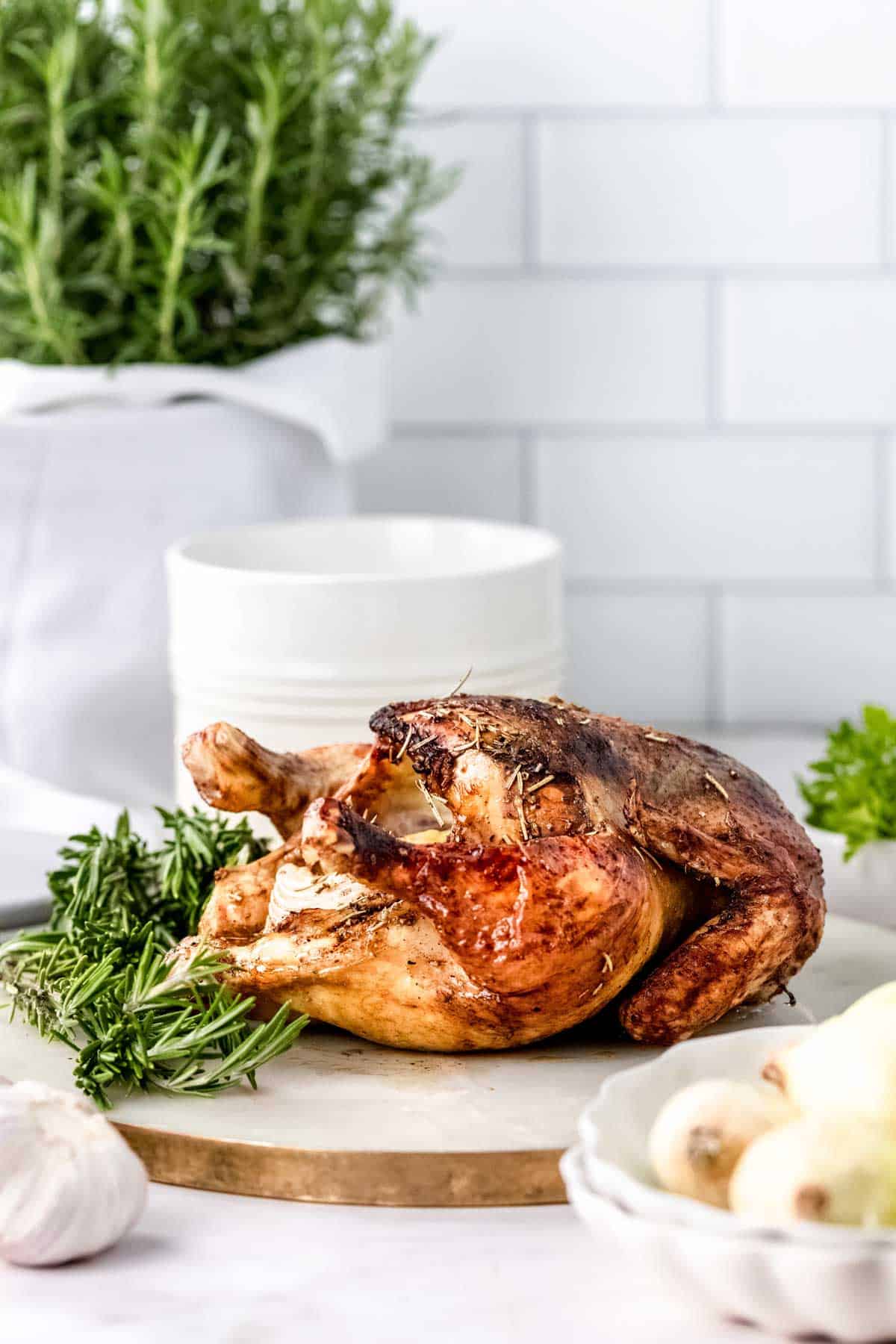 A whole rosemary balsamic roast chicken on a marble cutting board, next to a bowl of onions and fresh rosemary sprigs.