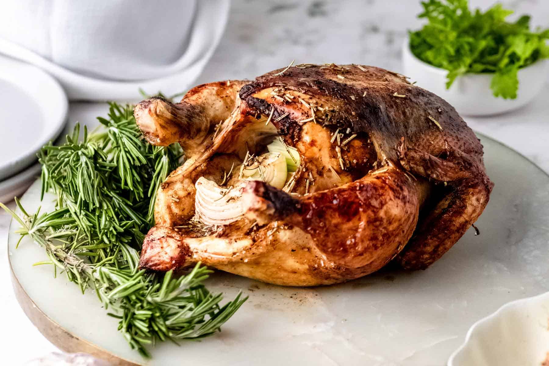 A whole rosemary balsamic roast chicken on a marble cutting board, next to fresh rosemary sprigs.