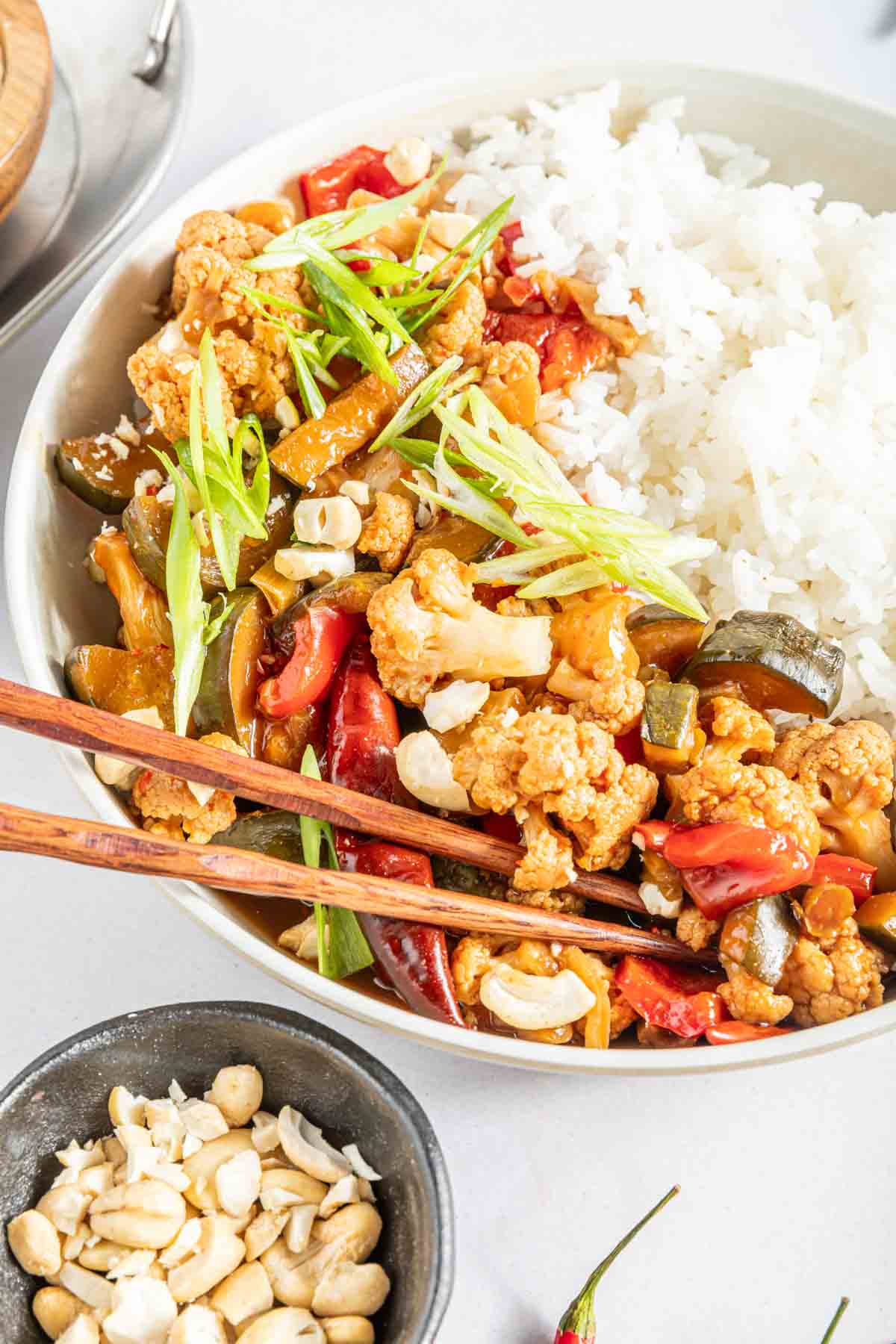 Kung pao cauliflower served in a bowl with white rice with chopsticks, next to a dish of crushed cashews.