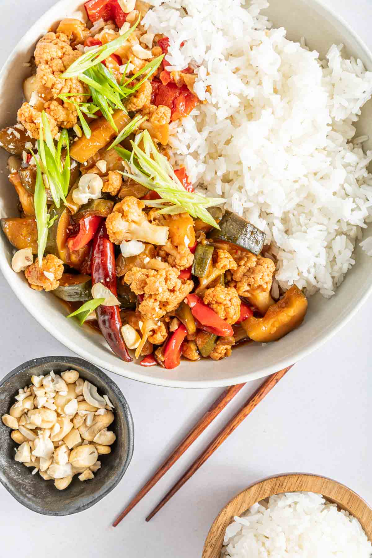 Kung pao cauliflower served in a bowl with white rice next to chopsticks and a dish with crushed peanuts.