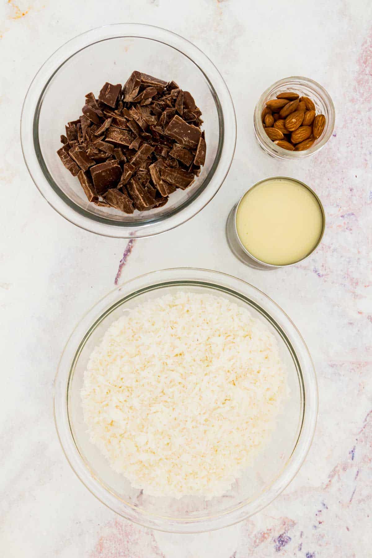 Bowls of shredded coconut, whole almonds, and chopped chocolate, and an open can of sweetened condensed milk on a marble countertop.