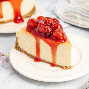 A slice of gluten free cheesecake on a plate with cherry topping and some of the cherry sauce dripping over the side onto the plate.