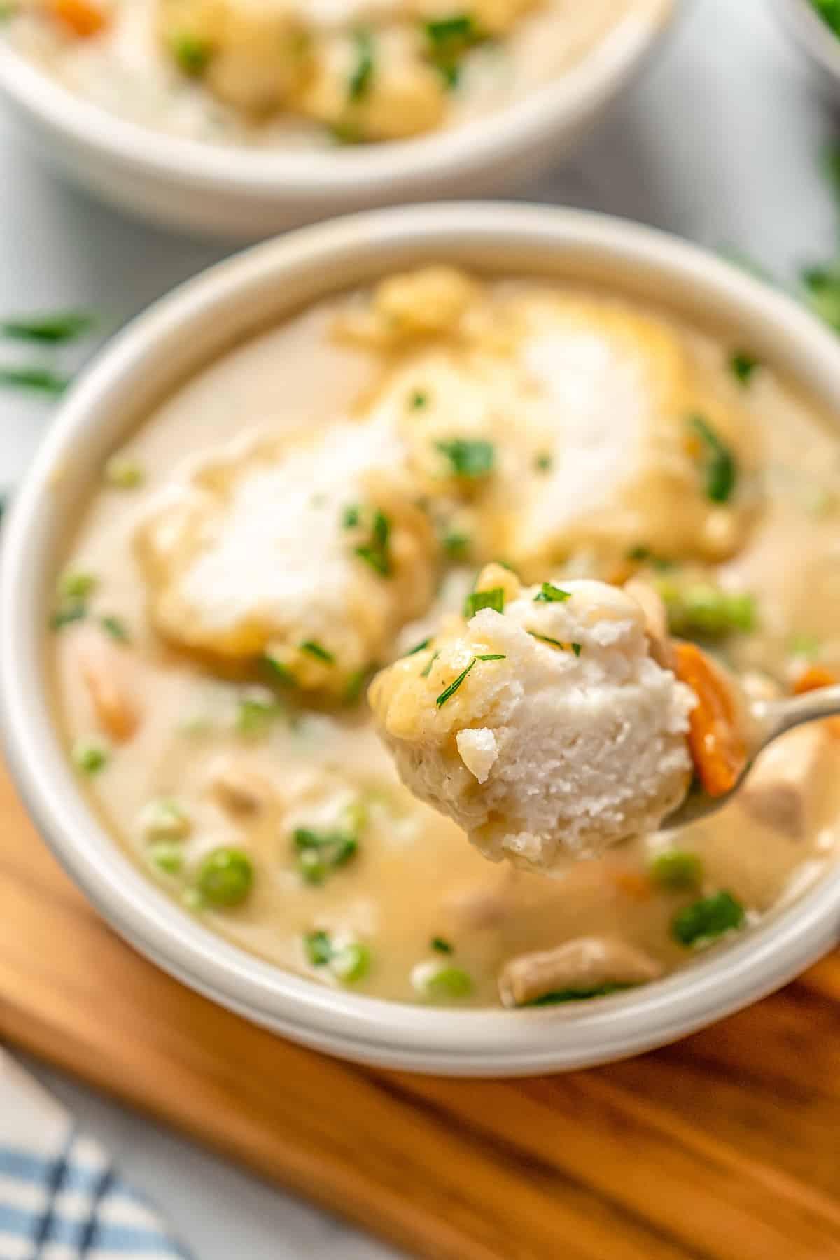 A dumpling on a spoon held in front of a bowl of gluten-free chicken and dumpling soup on a wooden board.