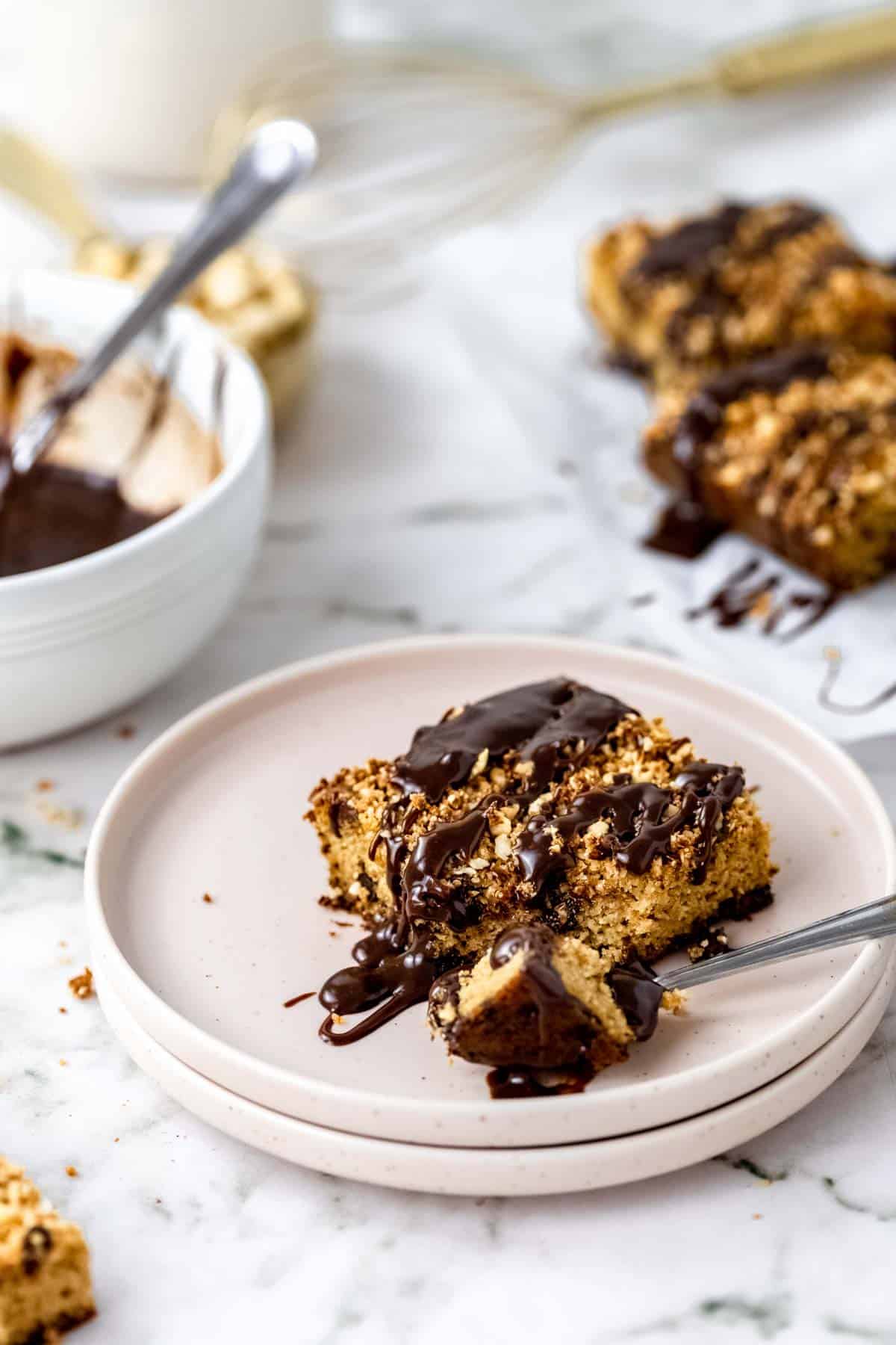 A slice of gluten-free chocolate hazelnut crumb cake on a plate, drizzled with chocolate glaze next to a fork.