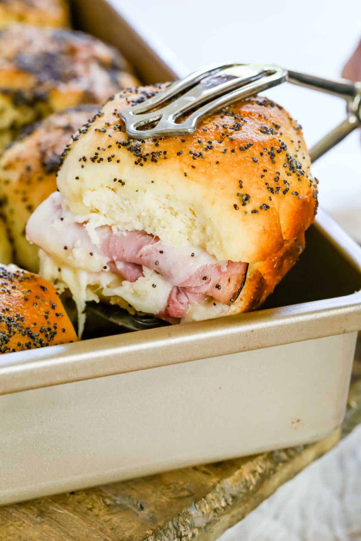 Tongs lifting a gluten-free ham and cheese slider from a baking pan.